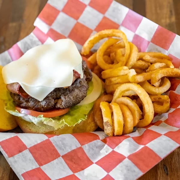 A cheeseburger with lettuce, tomato, onion, and bacon, served with curly fries in a red and white checkered basket.
